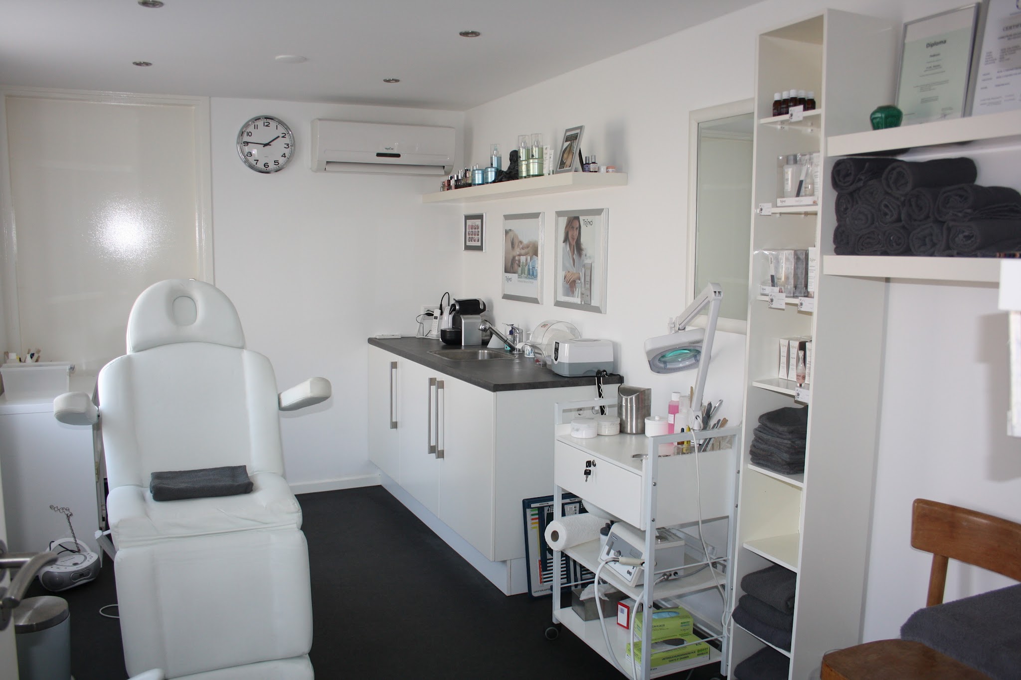 De lichte behandelruimte van Salon Vival met een witte behandelstoel, kasten en een klok aan de wand