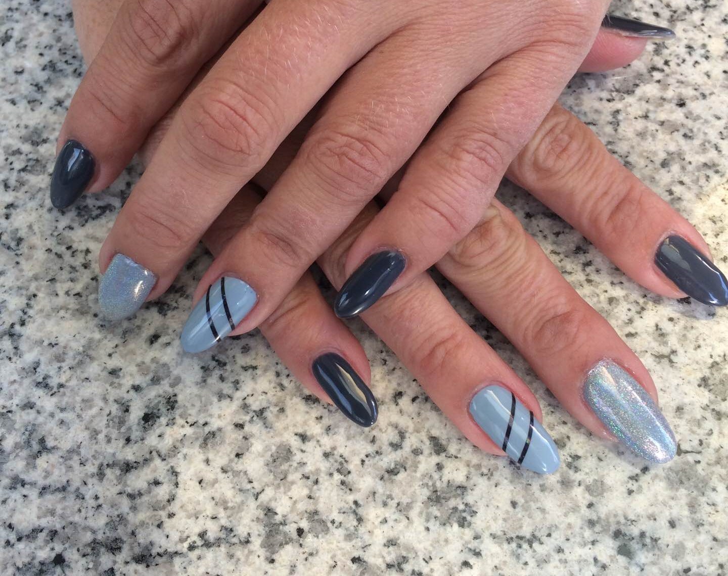 Twee handen met gelnagels in grijs en lichtblauw met dunne lijnen, op een granieten tafel