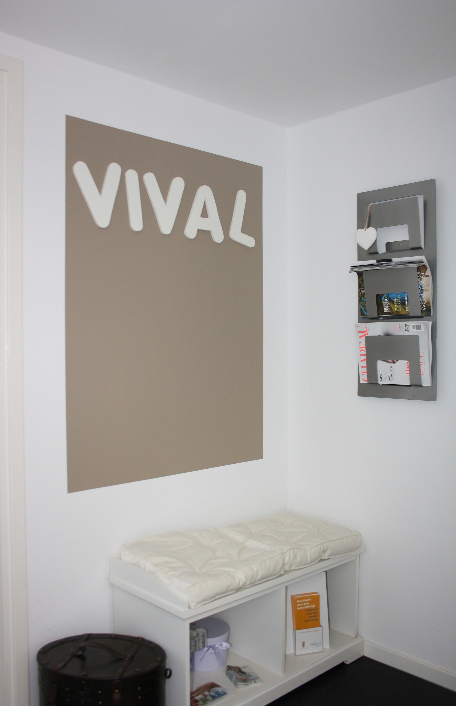 De taupe wand met de witte letters VIVAL en een wit bankje in de salon